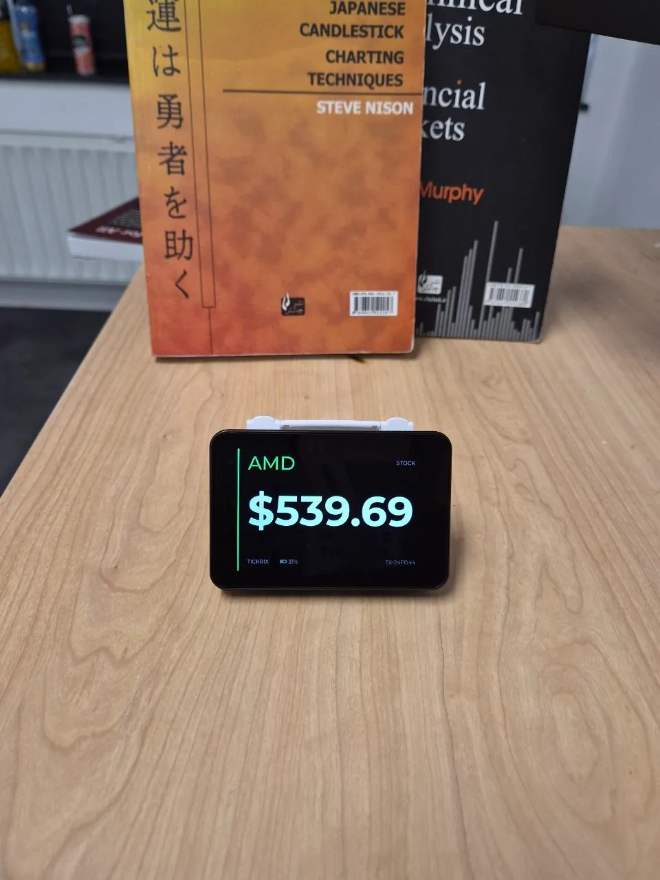 A Tickrix stock ticker display showing the AMD share price full-screen, on a desk in front of technical analysis books